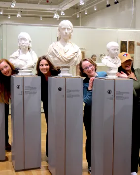 “A group of people playfully pose by leaning out between classical marble busts in a museum gallery.