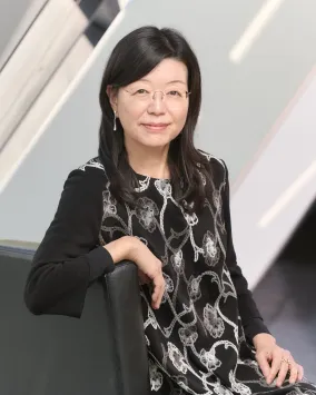 Asian woman with glasses wearing a black dress with a white floral pattern, sitting on a black chair against a modern background.