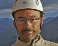 Man wearing glasses and a white Petzl helmet with a cloudy sky and mountains in the background.