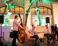 A three-member band performs inside a museum under a large dinosaur skeleton with green lighting.