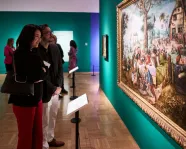 Two visitors closely observing a large framed painting displayed on a teal gallery wall.