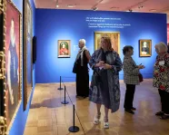 Five adults stand or walk inside an art gallery with blue and pink walls displaying framed paintings.