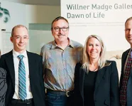 Five adults in business attire stand and smile in front of a sign reading “Willner Madge Gallery Dawn of Life.”