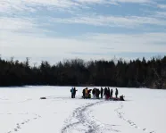 Group of people gathered on a snowy frozen lake surrounded by trees under a partly cloudy sky.