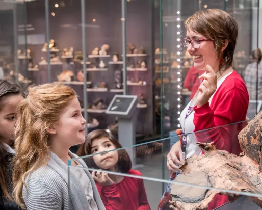 A woman wearing glasses and a red top talks to four children who are looking at artifacts behind glass in a museum.