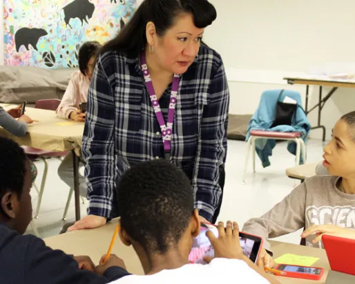 A teacher in a plaid shirt engages with four students seated around a table in a classroom.