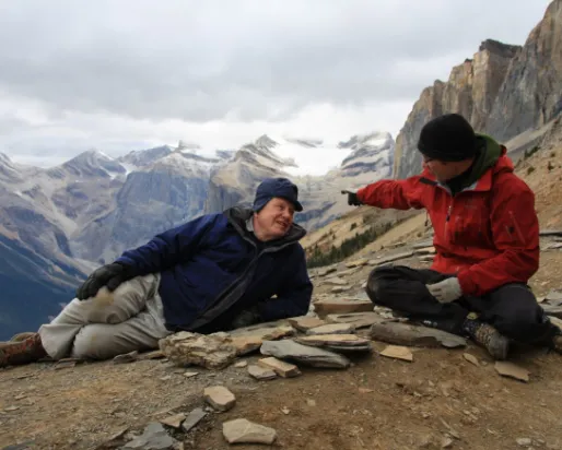 Two people sit and gesture on a rocky mountain trail with towering cliffs and distant peaks in the background.