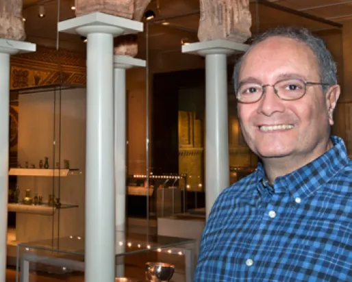 A man wearing glasses and a blue checked shirt stands in a museum gallery with white columns and display cases behind him.