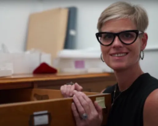 A smiling person with short blonde hair and black glasses holds a long thin wooden artifact in a workspace.