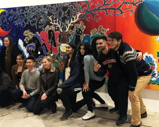 Group of young people posing and smiling in front of a colorful museum mural featuring abstract shapes and figures.