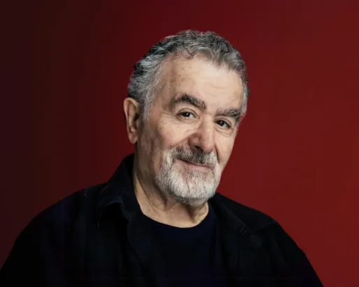 Portrait of an older man with gray hair and beard wearing a black shirt against a maroon background.
