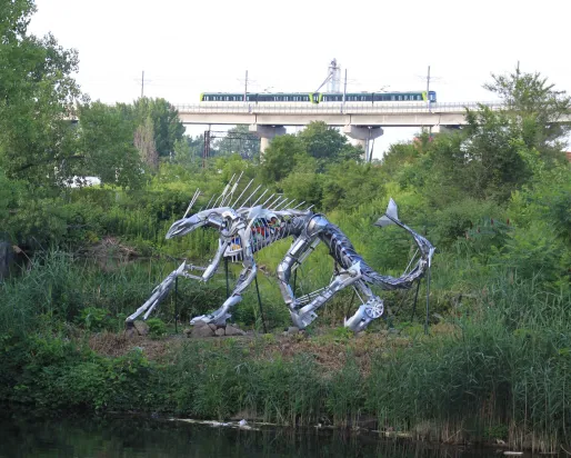 Steel sculpture of a crouching sabre-toothed cat surrounded by greenery near a body of water, with a passing train in the background.