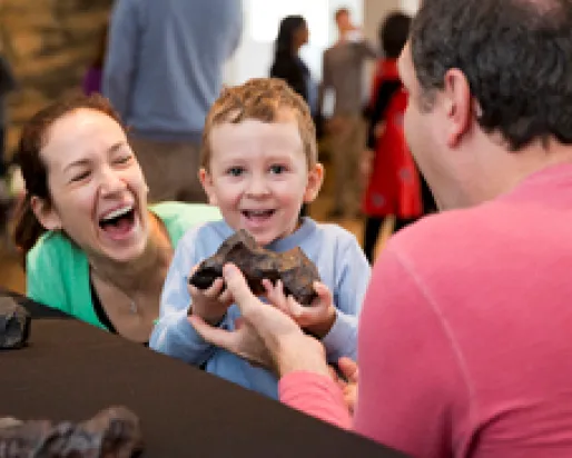 A smiling child with red hair holds a small artifact, with a laughing woman and a man looking on.