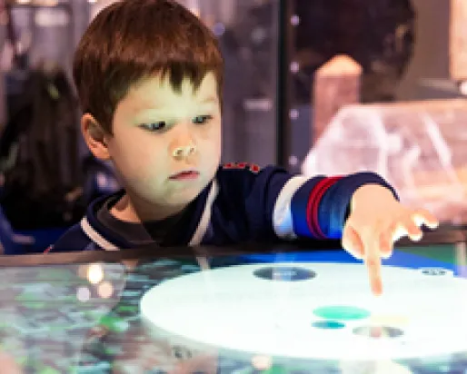 A young boy with dark hair interacts with a circular touchscreen display, with exhibit items blurred in the background.