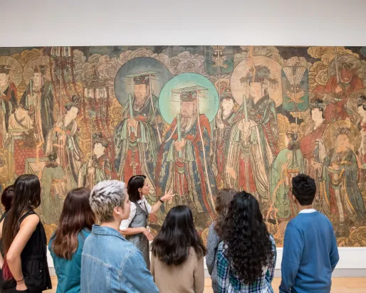 A group of students listens to a guide in front of a large traditional East Asian mural in a museum.