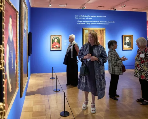 Five adults stand or walk inside an art gallery with blue and pink walls displaying framed paintings.