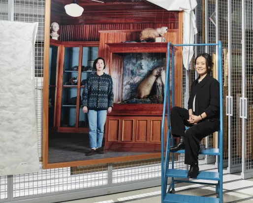 Two women pose near a large photographic print hanging from a metal grid; one woman stands in front while the other sits on a ladder.