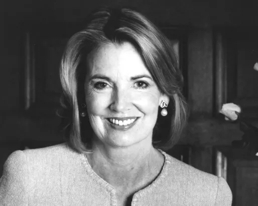 Portrait of a woman with light-colored hair, wearing a light jacket, smiling against a dark wooden background.