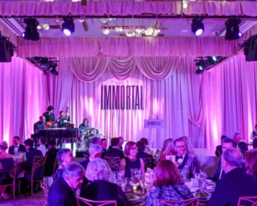 A banquet hall with round tables filled with people under purple lighting and a stage with musicians playing.