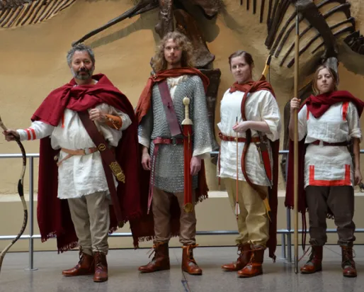 Four people dressed in ancient Roman soldier costumes standing indoors with a large dinosaur fossil mounted on a wall behind them.