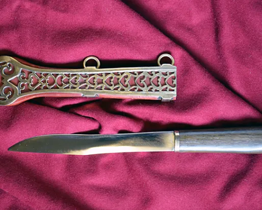 Steel knife with a straight blade and a polished metal handle next to an ornately decorated metal sheath on red cloth.