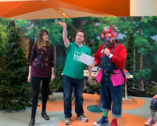 Three people stand on a colorful stage with a leafy green backdrop, one person in a red costume holds an item, and one person is seated on the right.