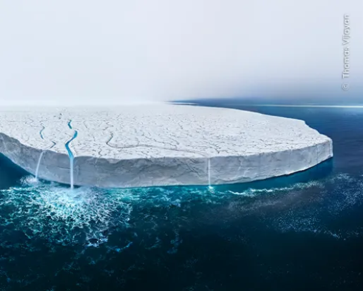 A large ice shelf floating on calm, dark blue ocean water under a clear sky.