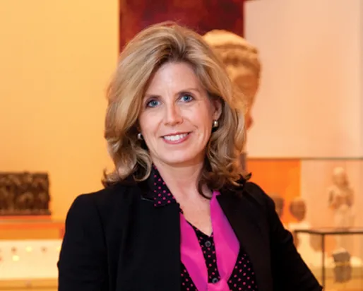 Woman with shoulder-length blonde hair wearing a black blazer over a pink blouse, standing indoors with blurred background.
