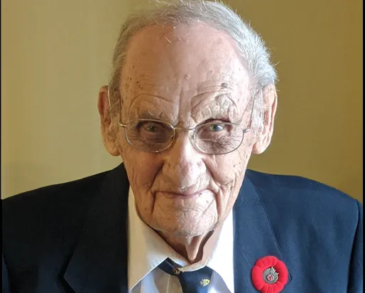 Elderly man wearing a dark blazer, striped shirt, and tie with a red poppy pin on his lapel.