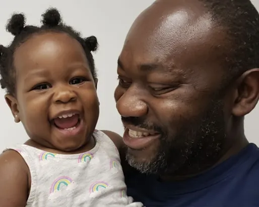 A smiling man in a blue shirt holding a laughing toddler wearing a white tank top with rainbow patterns.