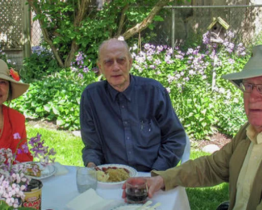 Three older adults sitting outdoors at a table with flowers and greenery in the background.