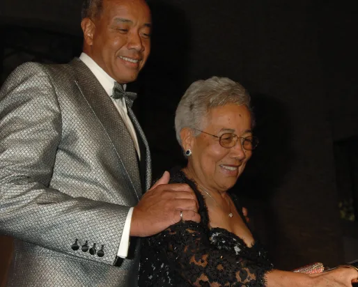 A man in a silver suit jacket stands behind a woman in glasses and a black lace dress.