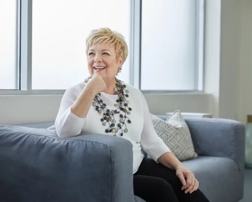 Portrait of Janice Price smiling and seated on a gray sofa, wearing a white blouse and a statement necklace.