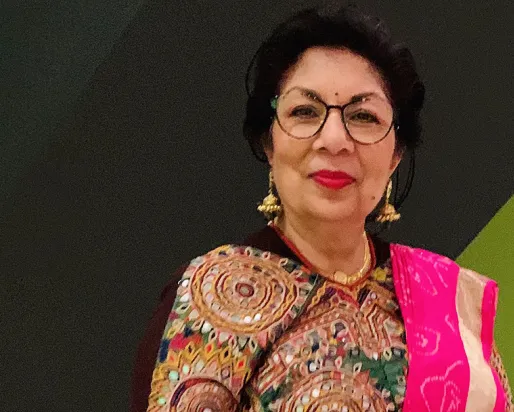 A woman wearing a brown patterned top, gold necklace, glasses, and a bright pink shawl on her left shoulder against a green and black background.
