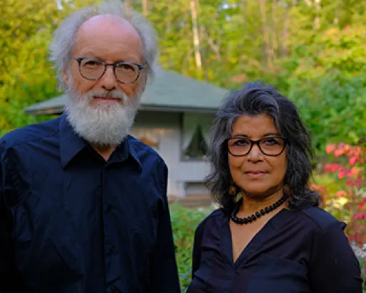 Portrait of an older man with white hair and beard wearing glasses and a black shirt, and a middle-aged woman with dark hair and glasses wearing a black top, standing outdoors with green trees and a pavilion in the background.