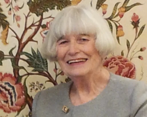 Elderly woman with white hair smiling in front of a floral patterned background.