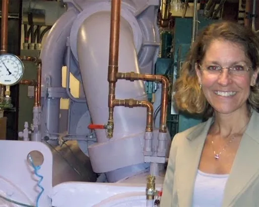 A woman with glasses and curly hair stands smiling in front of industrial pipes and gauges.