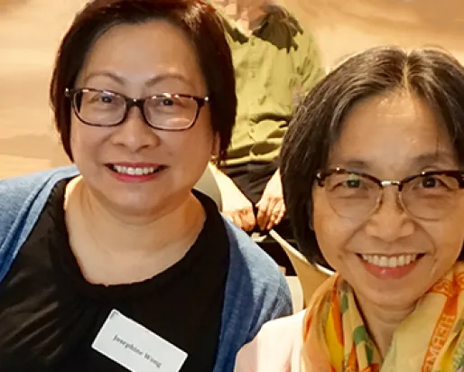 Two smiling women wearing glasses, seated indoors with one wearing a name tag.