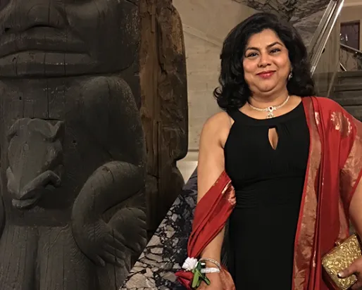 A woman in a black dress with a red shawl stands beside a large carved wooden totem pole inside a building.