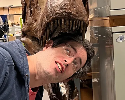 A person with dark hair leans their head toward the jaws of a large dinosaur skull mounted on a display stand.