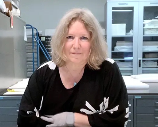 A woman with shoulder-length blonde hair wearing a black sweater with white skeleton designs sits at a desk in a laboratory or office setting.