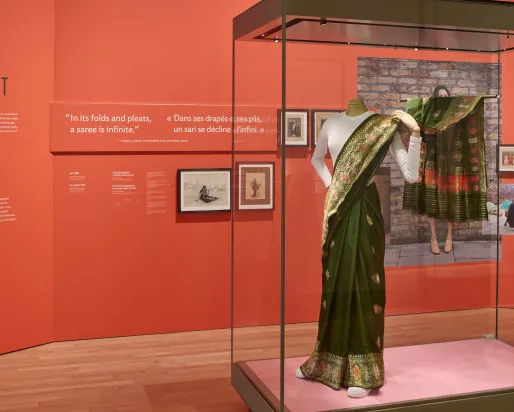Installation showing red text panels behind a glass case containing a green silk sari with woven metallic thread worn over a white T-shirt, shown on a mannequin wearing white running shoes.