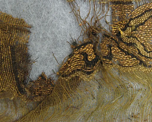 Fragment of ancient textile showing loose threads and woven fabric in brown and beige tones.