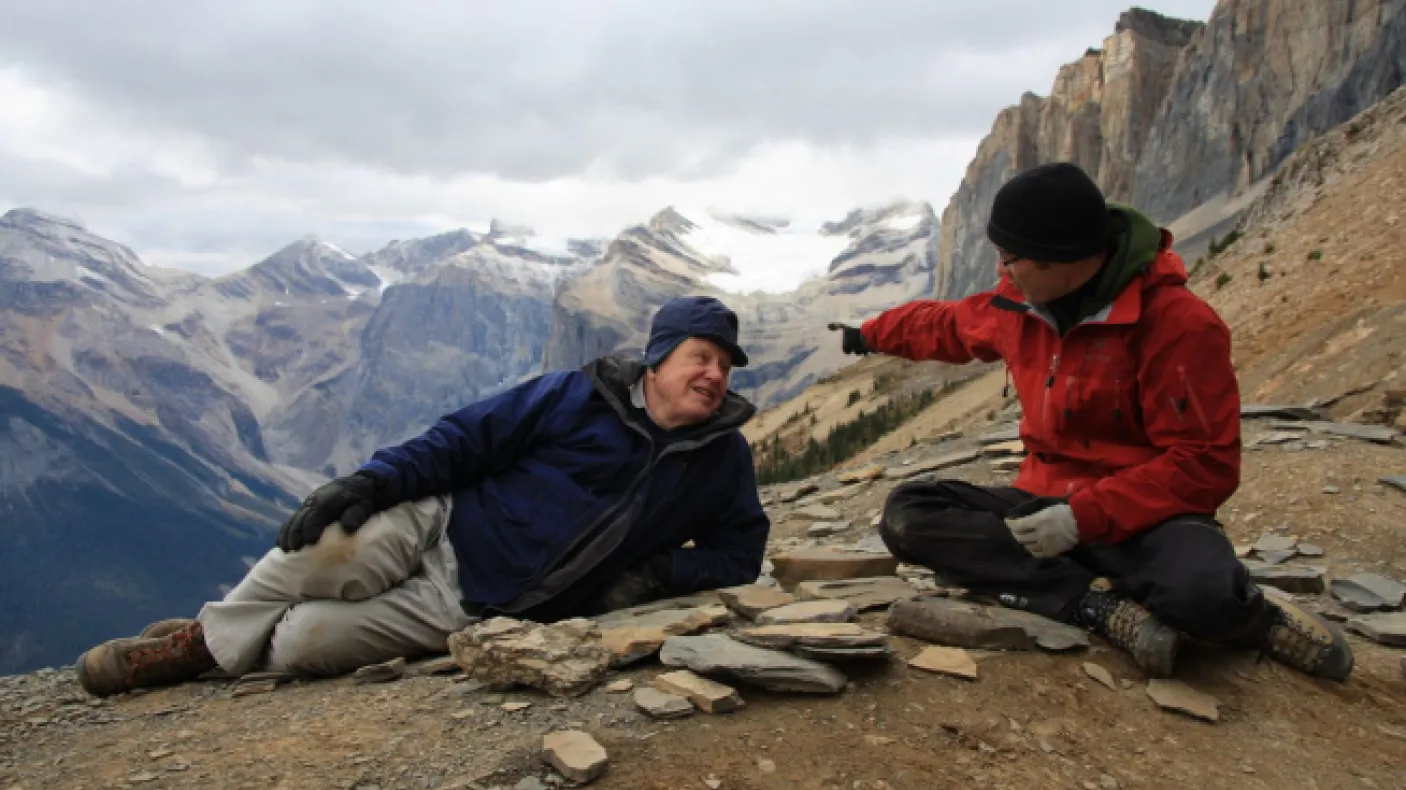 Two people sit and gesture on a rocky mountain trail with towering cliffs and distant peaks in the background.