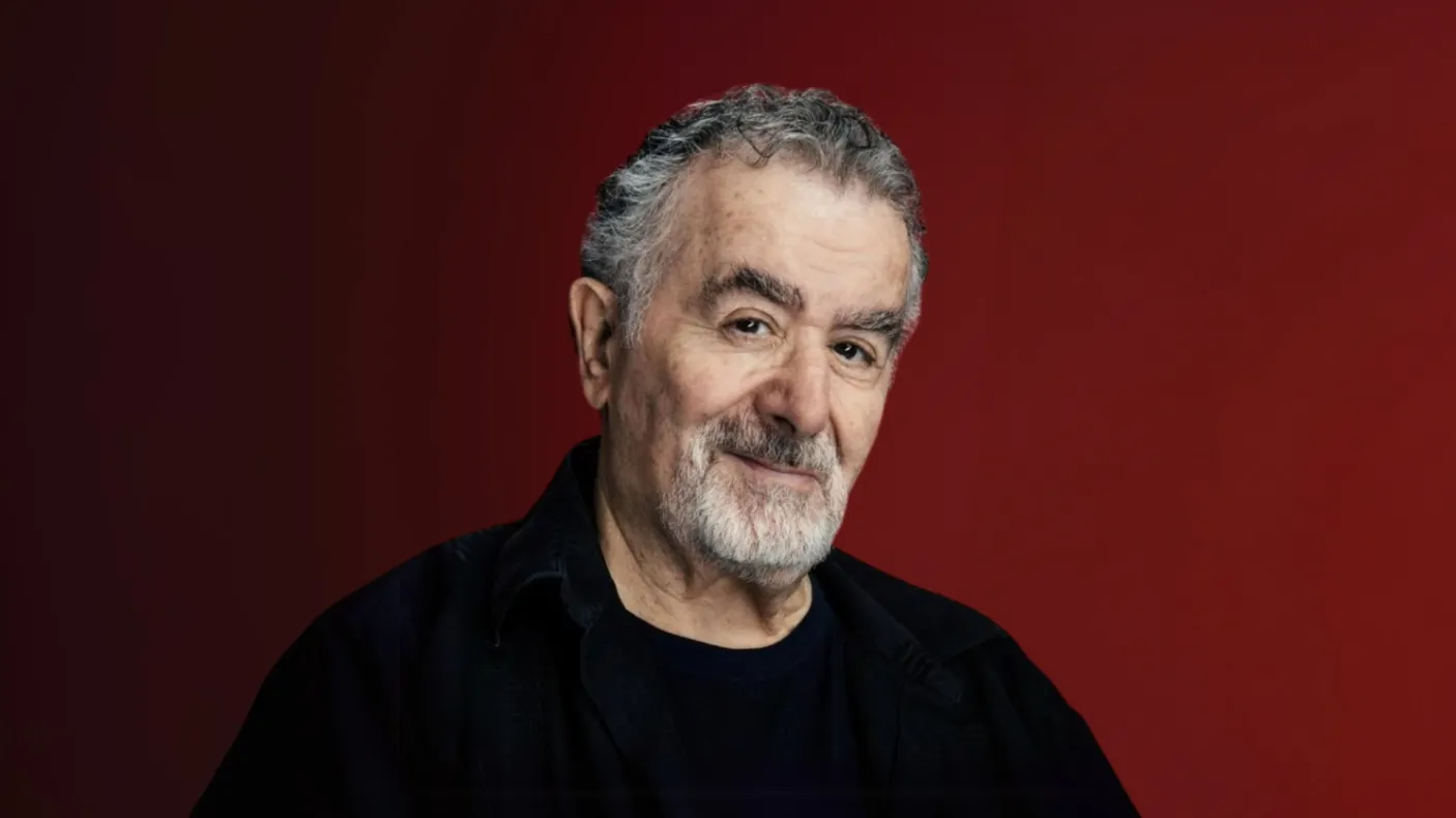 Portrait of an older man with gray hair and beard wearing a black shirt against a maroon background.