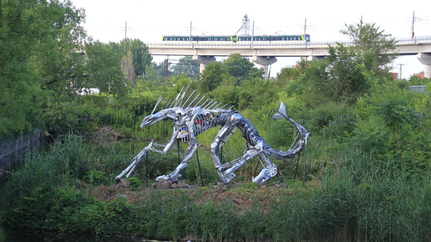 Steel sculpture of a crouching sabre-toothed cat surrounded by greenery near a body of water, with a passing train in the background.