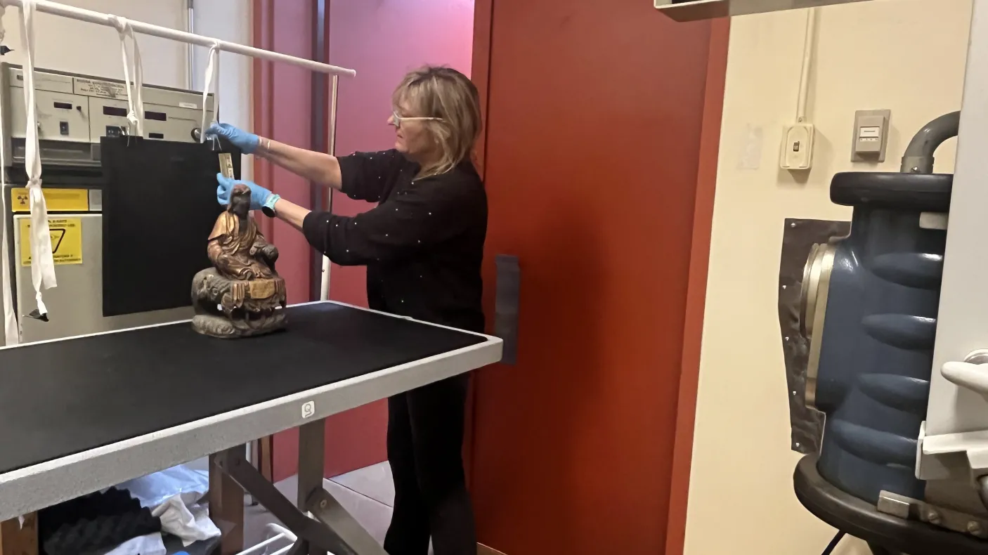 A woman adjusts a device near a Buddha statue on a metal table beside shelving and a red door.