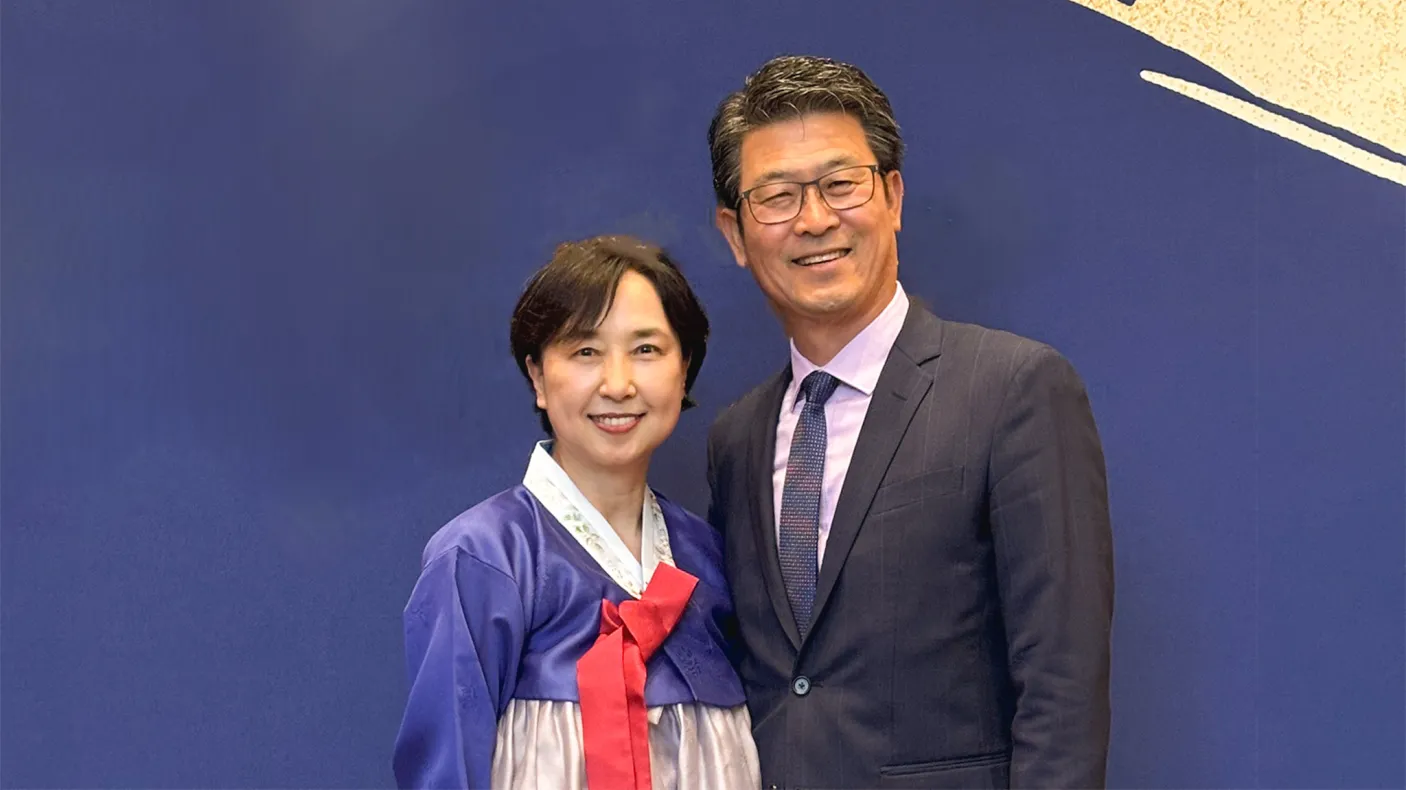 A man in a dark suit and glasses stands next to a woman wearing a traditional Korean hanbok dress with a blue top and red bow.
