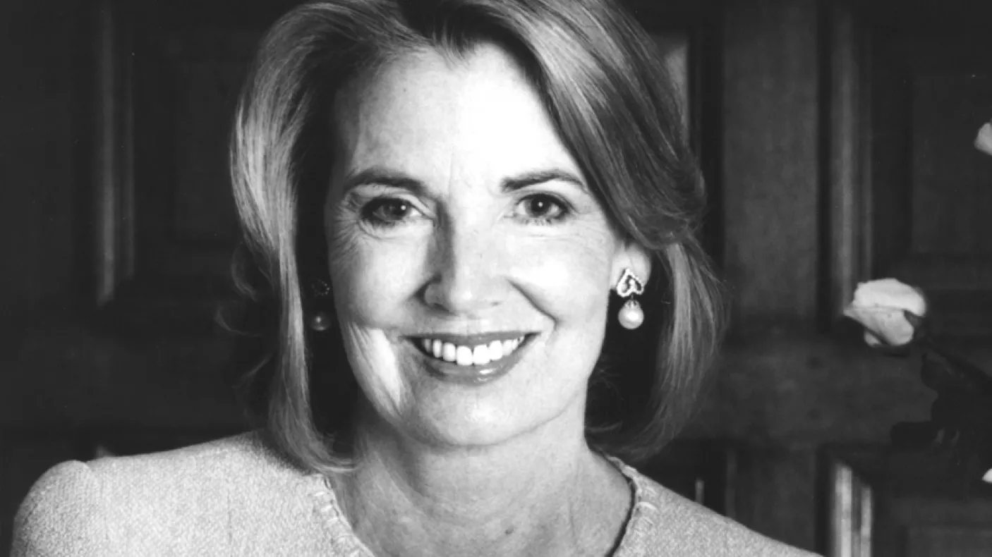 Portrait of a woman with light-colored hair, wearing a light jacket, smiling against a dark wooden background.