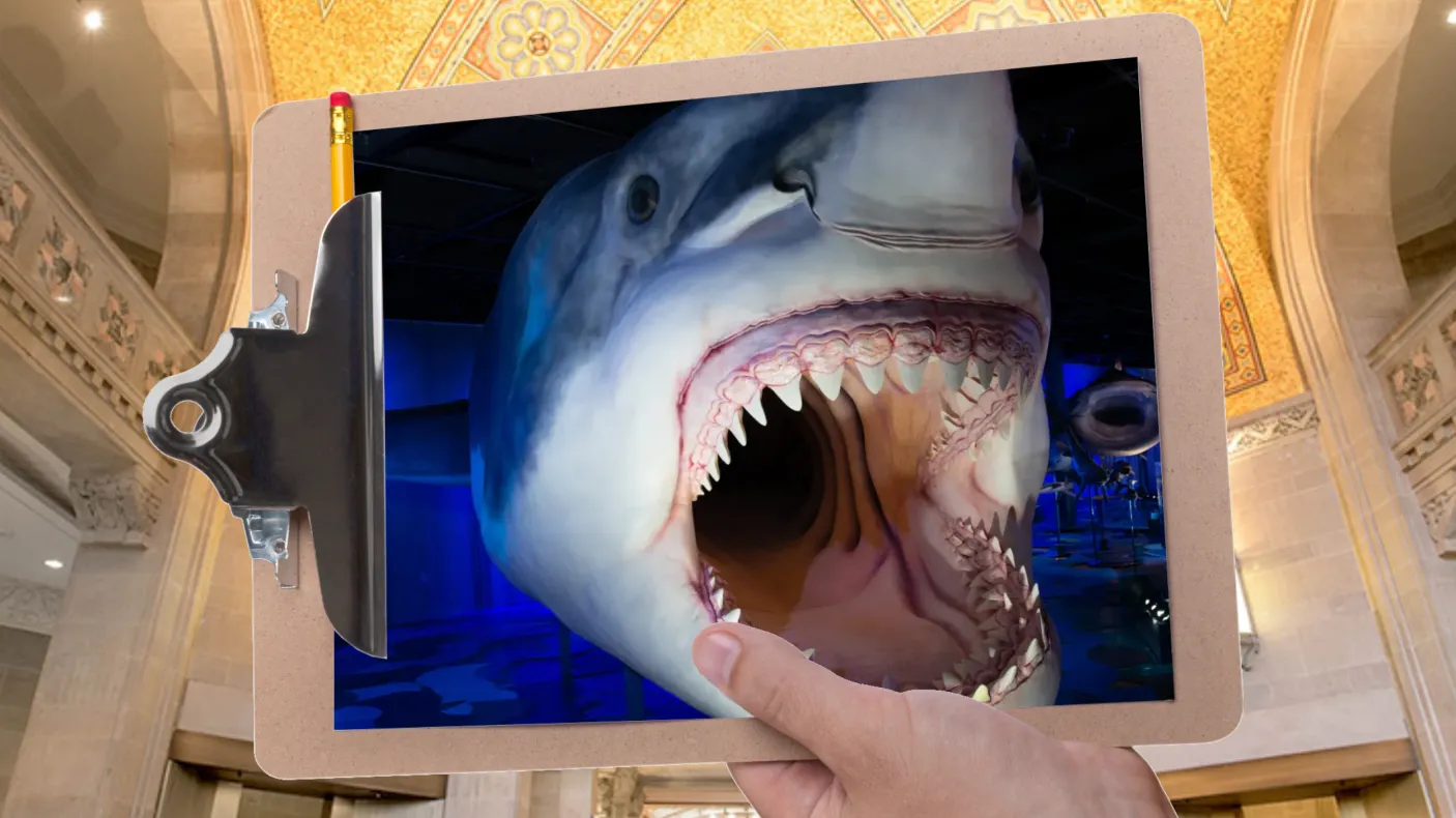 Une main tient un presse-papiers devant une galerie au plafond en mosaïque. Le presse-papiers contient la photo d'un énorme requin à la gueule béante.